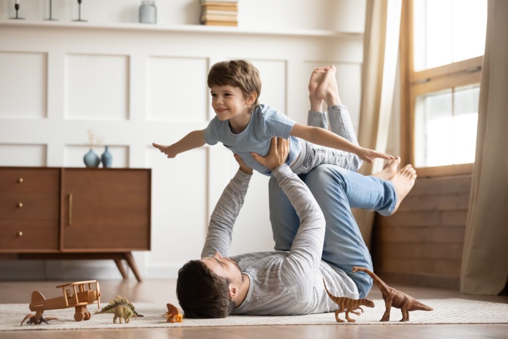 A father lies on his back, holding his smiling son aloft with his feet and hands as the boy pretends to fly.