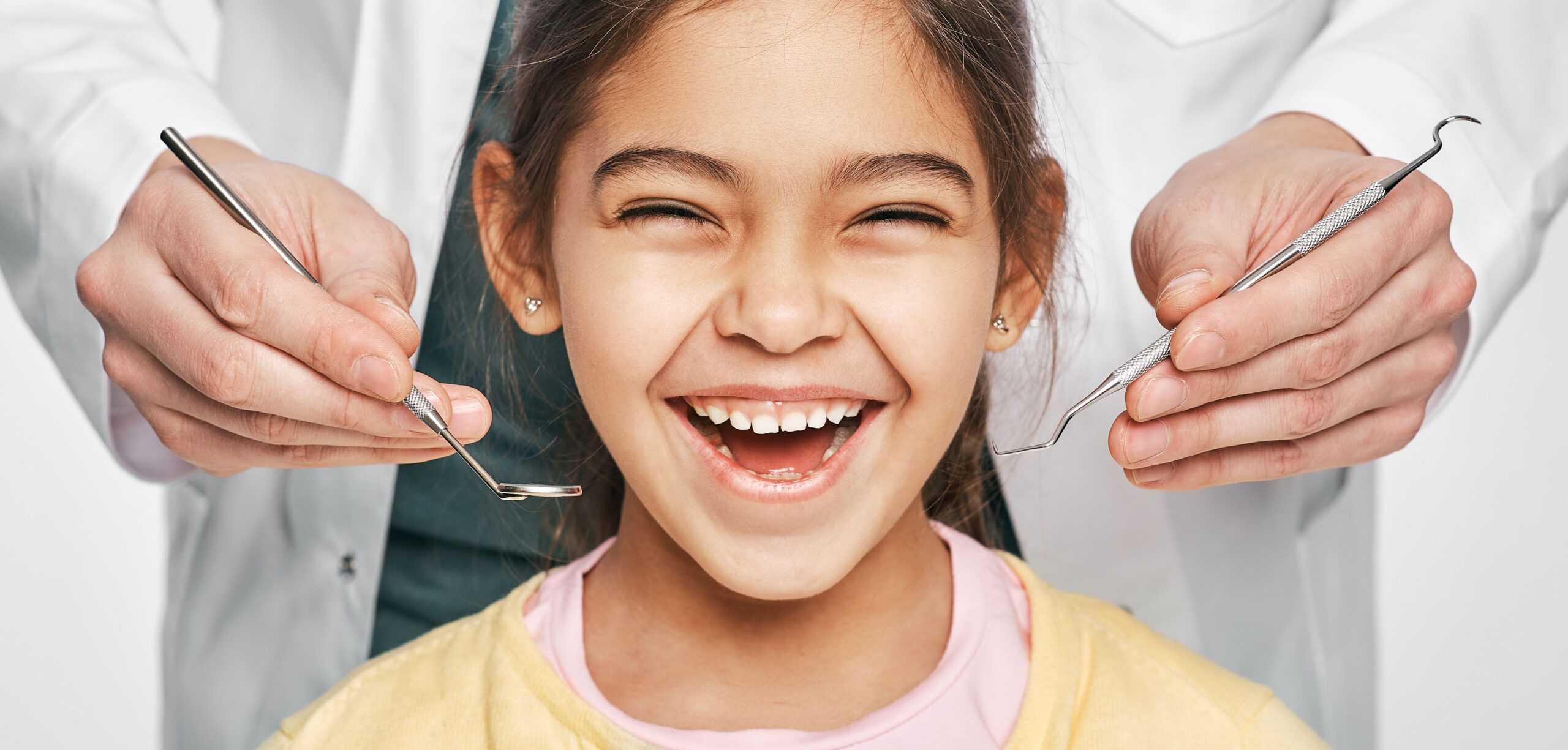 A happy young girl smiles widely as a dentist's hands hold dental tools.