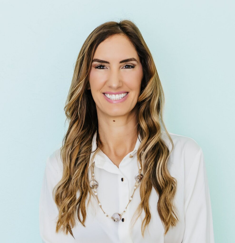 A smiling woman with long wavy hair, wearing a white shirt and necklace, against a light blue background.