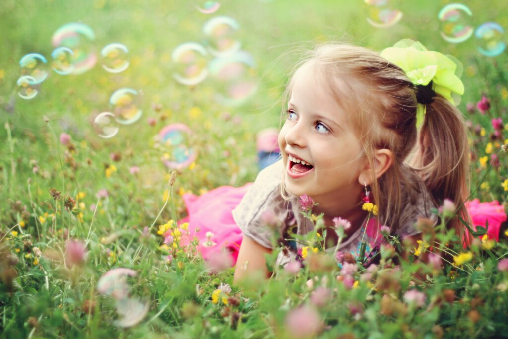 A smiling young girl lies in a grassy field with flowers, looking up at bubbles floating around her.