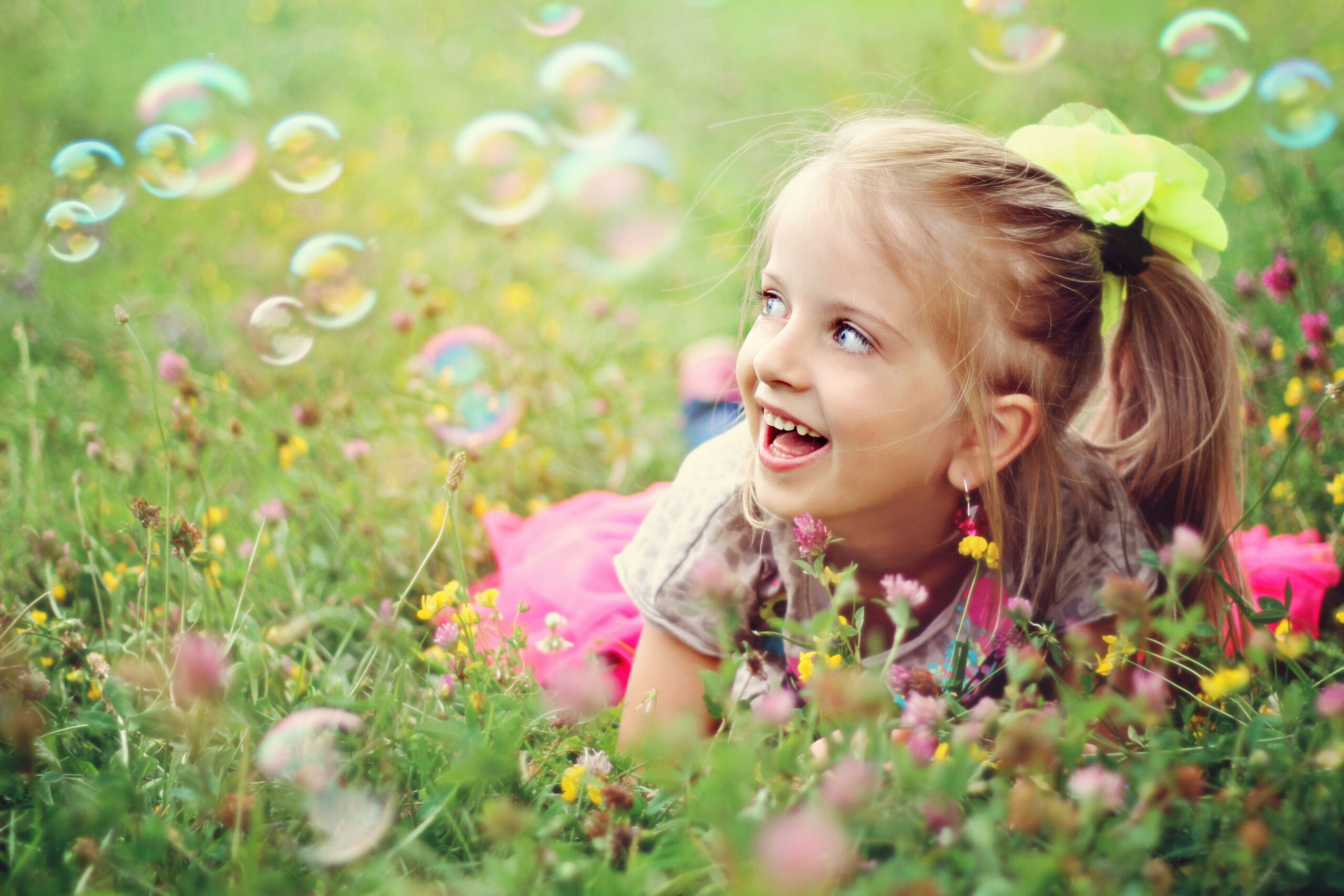 A smiling young girl lies in a grassy field with flowers, looking up at bubbles floating around her.