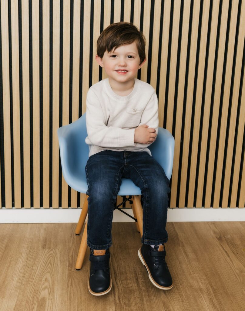 A young boy sits in a blue chair with his arms crossed, smiling at the camera, against a vertically striped wooden wall.