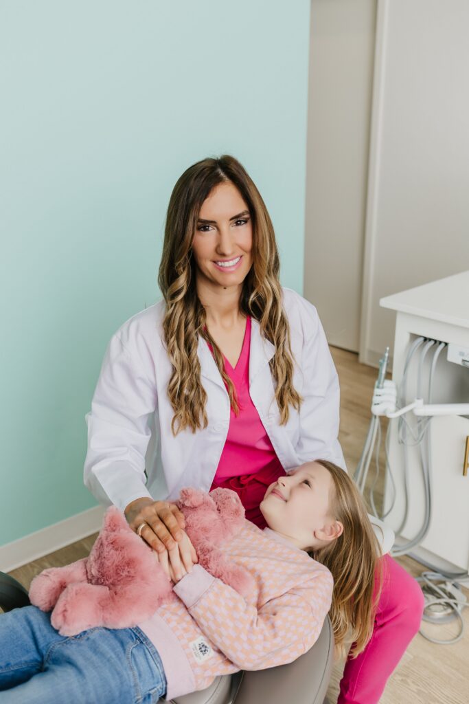 Female dentist smiling while comforting a young girl holding a pink stuffed animal in a dental chair.