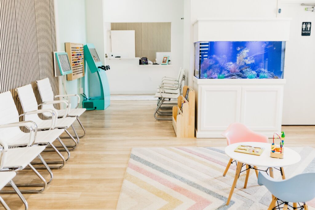 Modern waiting room with white chairs, a large aquarium, a children's play area with toys, and a reception counter.