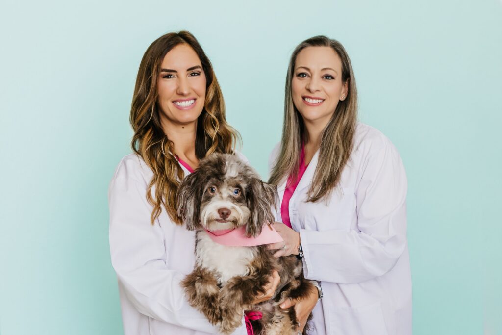 Two smiling women in lab coats and pink scrubs hold a fluffy dog with a pink bandana.