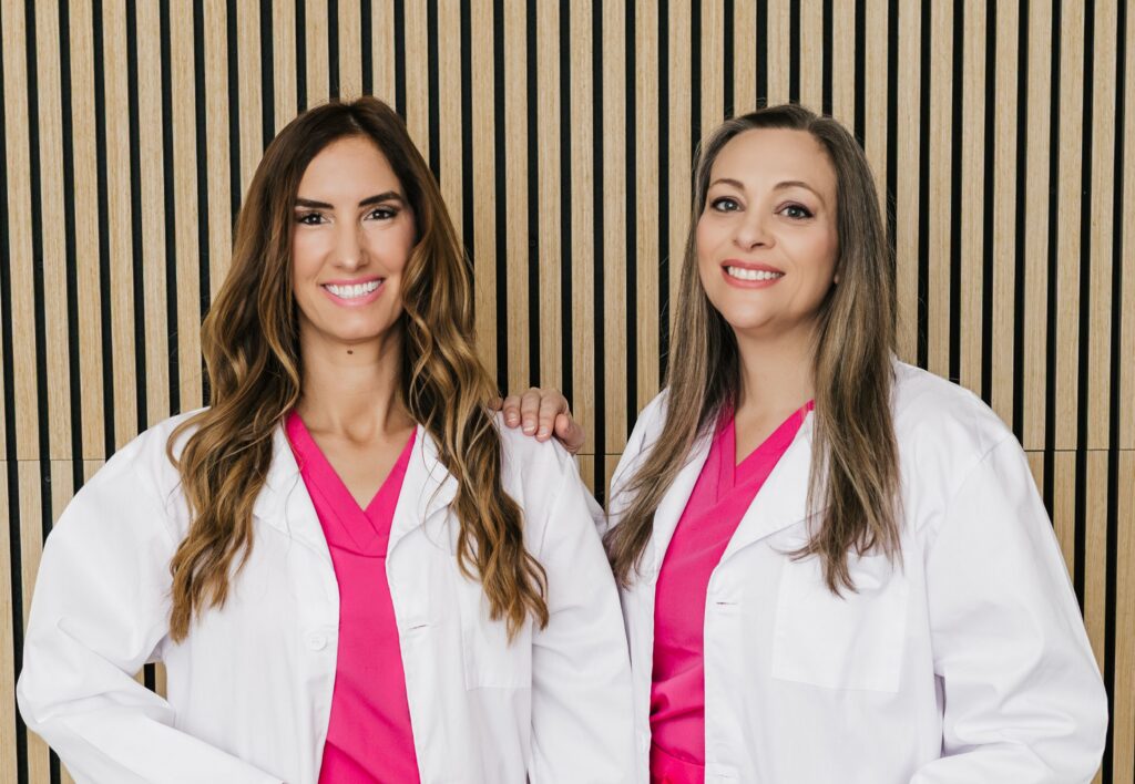 Two smiling women in white lab coats and pink scrubs stand against a striped wooden wall.