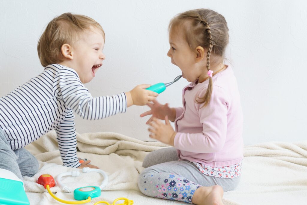 Two toddlers laughing while playing doctor with a toy dental tool.