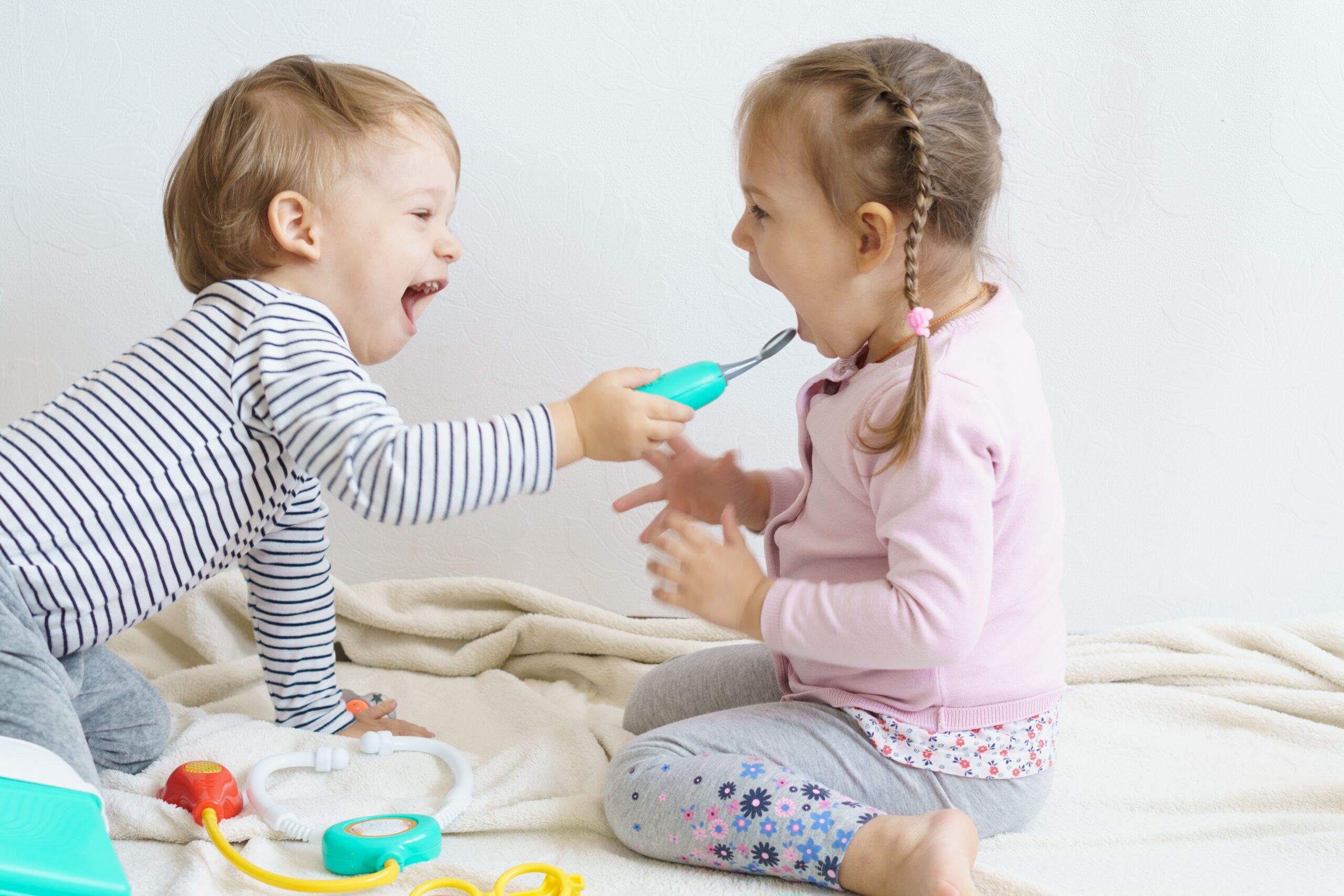 Two toddlers laughing while playing doctor with a toy dental tool.
