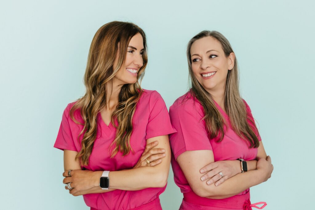 Two women in pink scrubs smile at each other, standing back-to-back with arms crossed.
